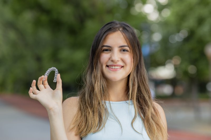 a young woman holding a clear Invisalign aligner in her right hand and smiling